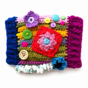 May include: A colorful, handmade crochet item with a variety of textures and embellishments. The piece features a patchwork design with blue, yellow, green, and purple sections. It is adorned with crochet flowers, buttons, and a square floral motif.