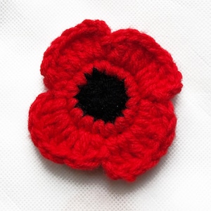 May include: A close-up of a crocheted poppy, a symbol of remembrance, in bright red with a black central disc. The handmade flower features detailed stitching, showcasing the texture of the yarn. A classic floral design.
