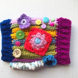 May include: A colorful, handmade crochet item with a patchwork design. It features a red square with a flower, a purple flower, and various buttons. The edges are blue and purple, with a white and gray trim.