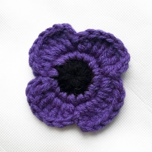 May include: A close-up of a handmade crochet flower. The flower is predominantly purple with a black centre. The petals are textured and detailed, suggesting a handcrafted item. The flower is likely intended for decorative purposes or as an embellishment.