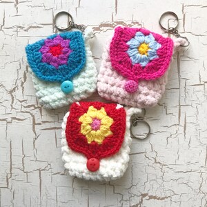 May include: Three handmade crochet backpack keychains in pastel colors. Each keychain features a floral design on the flap and a button closure. The keychains are in blue, pink, and white, with silver key rings.