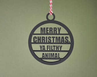 Op de afbeelding: Zwarte kerstbal met de tekst "MERRY CHRISTMAS, YA FILTHY ANIMAL" uitgesneden. De ronde versiering heeft een lus aan de bovenkant om op te hangen. Het hangt aan een rood-wit gestreept touwtje.
