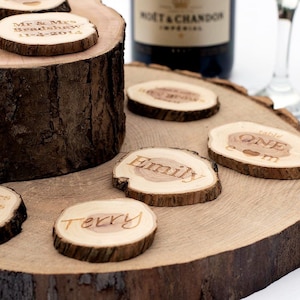 May include: Wooden place cards with engraved names and dates, displayed on a rustic wooden slab. The cards feature names like "Emily" and "Terry", and the words "Mr & Mrs Bradshaw 11-4-2014". A bottle of champagne is in the background.