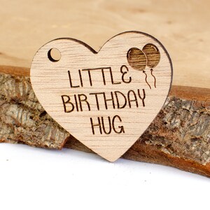 Peut inclure: Une étiquette en bois en forme de cœur avec le texte "Little Birthday Hug" et deux ballons avec des ficelles.