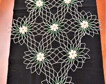 May include: A black table runner featuring a white, openwork floral design. The design consists of interconnected flower-like shapes with green and beige accents. The runner is placed on a wooden table.