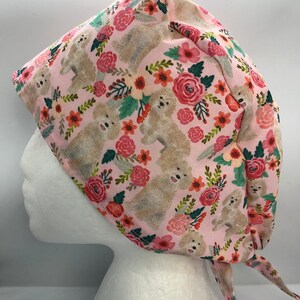 May include: A pink surgical scrub cap featuring a repeating pattern of light brown dogs and colorful flowers. The floral elements include red, pink, and orange roses, with green leaves. The cap is tied at the back.