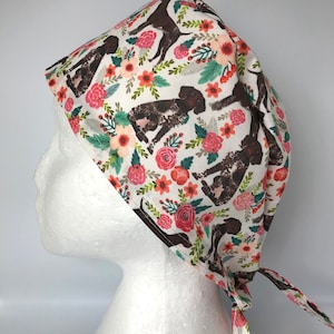 May include: A white scrub cap with a repeating pattern of brown and white dogs and colourful flowers. The flowers are in shades of pink, orange, and green. The cap is tied at the back.