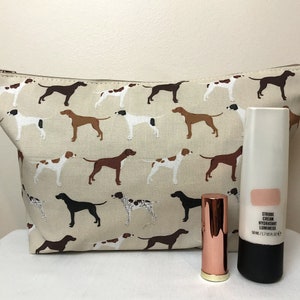 May include: A beige cosmetic bag featuring a pattern of dogs in brown, black, and white. Beside the bag are a tube of face cream and a rose gold lipstick.