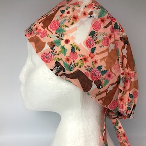 May include: A peach-coloured surgical scrub cap with a repeating pattern of boxer dogs, pink roses, and green foliage. The cap has a tie closure at the back and is designed to cover the head and secure hair.