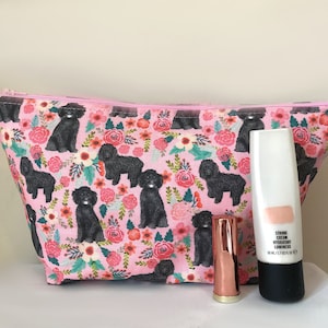 May include: A pink cosmetic bag featuring a pattern of black dogs and flowers, alongside a tube of strobe cream and a lipstick. The bag is zipped and contains beauty products.