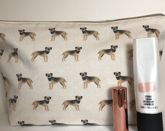 May include: A cream-coloured cosmetic bag featuring a repeating pattern of brown and black dogs. Beside the bag are a rose gold lipstick tube and a white tube of strobe cream. The bag has a zip closure.