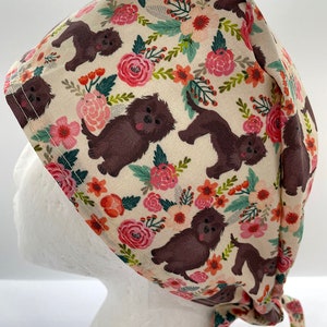 May include: A beige surgical scrub cap featuring a repeating pattern of brown dogs and pink, orange, and red flowers. The cap is tied at the back with matching fabric ties. The cap is designed to cover the hair during medical procedures.