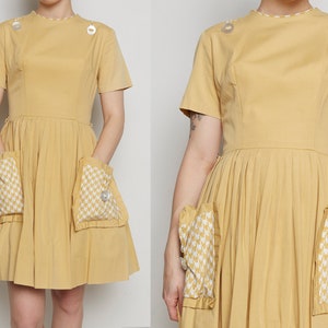 May include: A yellow vintage dress with short sleeves and a pleated skirt. The dress has two large pockets with a white and black checkered pattern and a button on each pocket.
