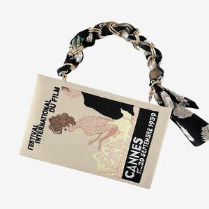 Könnte beinhalten: Eine beige Clutch mit einem schwarzen und goldenen Kettenriemen und einem schwarz-weißen Blumendruck. Die Tasche zeigt ein gesticktes Bild einer Frau in einem Kleid und den Text "Festival International du Film Cannes 1-20 Septembre 1959".