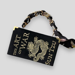 K&ouml;nnte beinhalten: Eine schwarze Buch-Tasche mit einem goldenen Kettengriff und einem schwarzen und wei&szlig;en Blumenmuster-Riemen. Die Buch-Tasche ist mit einer goldenen Stickerei eines Drachen und dem Text "The Art of War Sun Tzu" verziert.