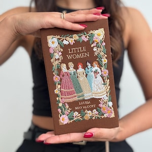 Op de afbeelding: Een bruine, geborduurde boekclutch met de titel "Little Women" van Louisa May Alcott. De omslag heeft een bloemmotief en een illustratie van vier vrouwen in jurken.