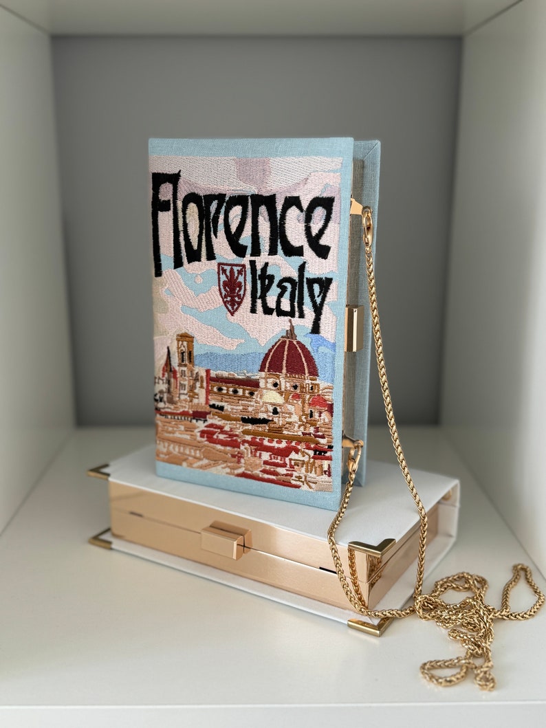 Pu&ograve; includere: Una pochette blu chiaro con una tracolla a catena dorata. La borsa &egrave; ricamata con una scena di Firenze, Italia, e il testo "Florence Italy".