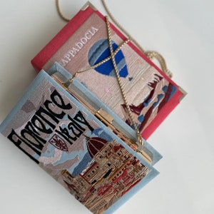 Pu&ograve; includere: Due pochette ricamate con catene. La borsa superiore &egrave; rossa con una mongolfiera e il testo "Cappadocia". La borsa inferiore &egrave; blu con una vista panoramica della citt&agrave; e il testo "Florence Italy".