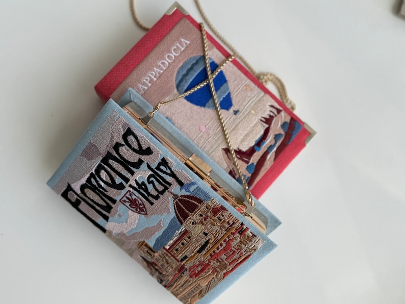 Pu&ograve; includere: Due pochette ricamate con catene. La borsa superiore &egrave; rossa con una mongolfiera e il testo "Cappadocia". La borsa inferiore &egrave; blu con una vista panoramica della citt&agrave; e il testo "Florence Italy".