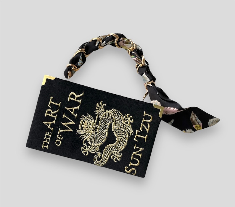 K&ouml;nnte beinhalten: Eine schwarze Buch-Tasche mit einem goldenen Kettengriff und einem schwarzen und wei&szlig;en Blumenmuster-Riemen. Die Buch-Tasche ist mit einer goldenen Stickerei eines Drachen und dem Text "The Art of War Sun Tzu" verziert.