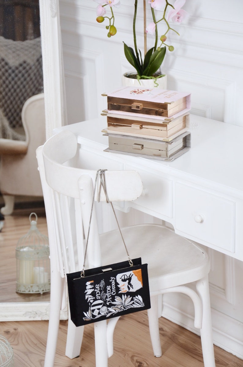 May include: A black clutch bag with a silver chain strap. The bag is decorated with an embroidered image of a book cover with the title "Dante's Divine Comedy".