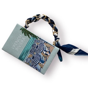 May include: A light blue clutch bag with a white embroidered design of beach umbrellas and the text "French Riviera". The bag has a gold chain strap with a blue and white patterned scarf attached.