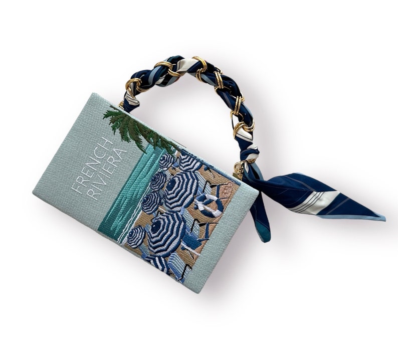 May include: A light blue clutch bag with a white embroidered design of beach umbrellas and the text "French Riviera". The bag has a gold chain strap with a blue and white patterned scarf attached.