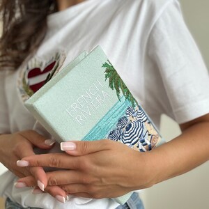 May include: A light blue book with the text "French Riviera" embroidered on the cover. The book features a beach scene with palm trees and umbrellas.