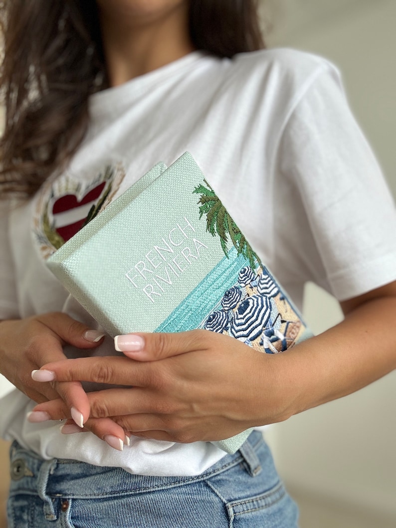 May include: A light blue book with the text "French Riviera" embroidered on the cover. The book features a beach scene with palm trees and umbrellas.