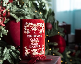 Könnte beinhalten: Ein rotes Samtbuch mit dem Titel "A Christmas Carol" von Charles Dickens, mit goldenen, verzierten Details, liegt auf einem verpackten Geschenk. Das Buch ist der Mittelpunkt, vor einem Weihnachtsbaum mit roten Ornamenten und Lichtern.