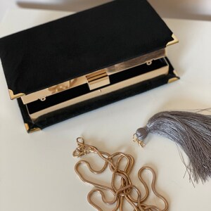 May include: A black velvet clutch with a gold clasp and chain strap. The clutch has a gray tassel.
