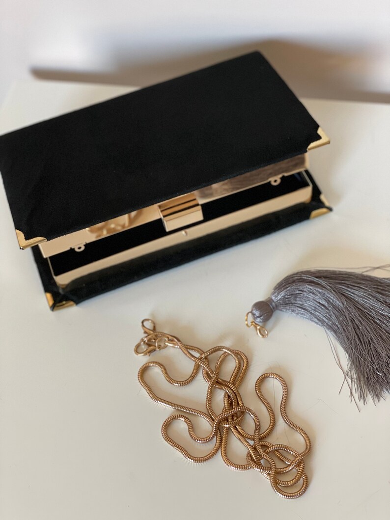 May include: A black velvet clutch with a gold clasp and chain strap. The clutch has a gray tassel.