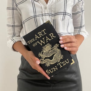 K&ouml;nnte beinhalten: Ein schwarzes Buch mit goldenen Lettern, auf dem "The Art of War" und "Sun Tzu" steht. Das Buch zeigt ein golden besticktes Drachenmotiv.