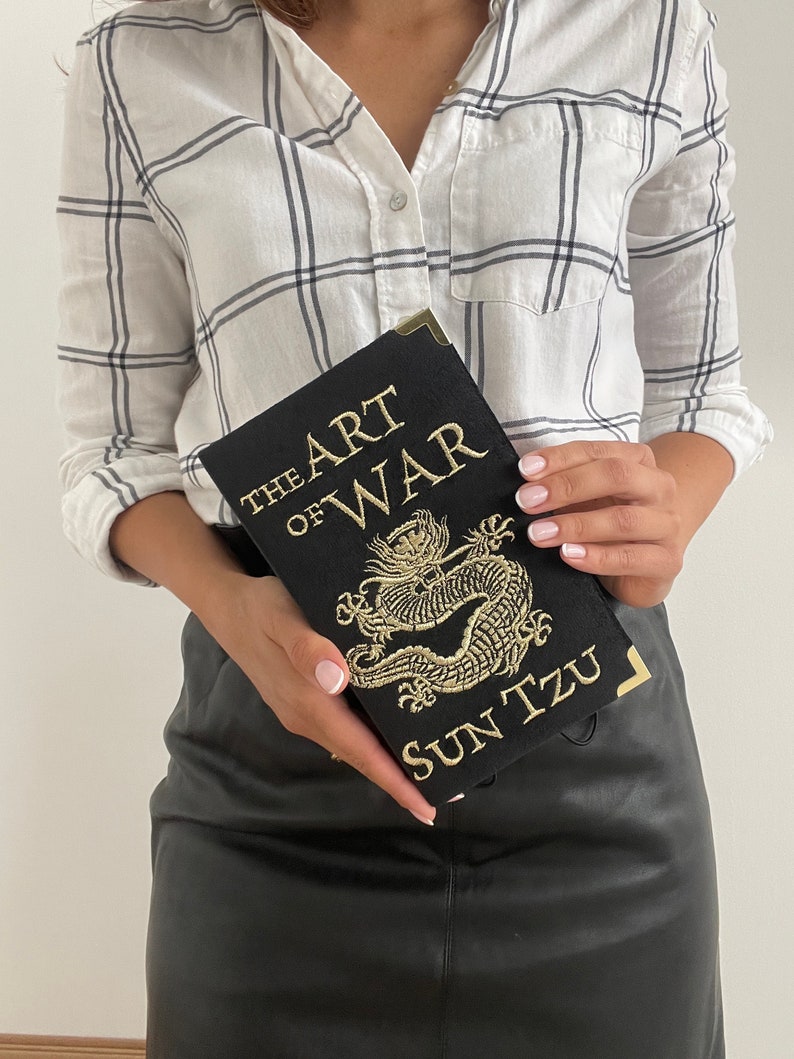 K&ouml;nnte beinhalten: Ein schwarzes Buch mit goldenen Lettern, auf dem "The Art of War" und "Sun Tzu" steht. Das Buch zeigt ein golden besticktes Drachenmotiv.