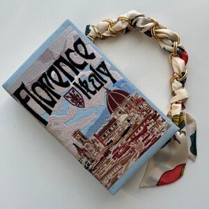 Pu&ograve; includere: Una pochette blu chiaro con una tracolla a catena dorata e una stampa floreale. La borsa presenta un'immagine ricamata del Duomo di Firenze, Italia, con il testo "Florence Italy" sopra l'immagine.