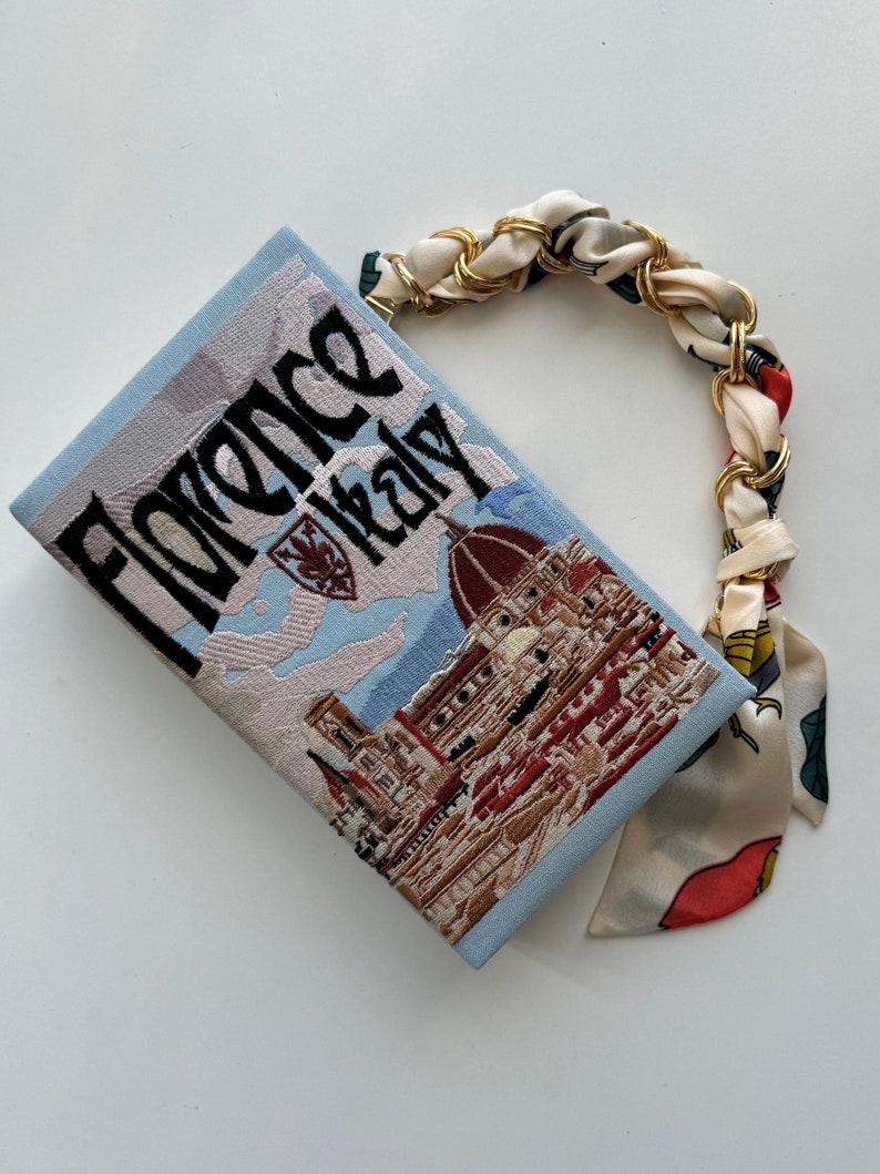 Pu&ograve; includere: Una pochette blu chiaro con una tracolla a catena dorata e una stampa floreale. La borsa presenta un'immagine ricamata del Duomo di Firenze, Italia, con il testo "Florence Italy" sopra l'immagine.