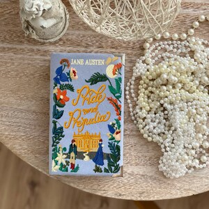 May include: A blue book cover with the title "Pride and Prejudice" embroidered in yellow and orange thread. The cover also features a small embroidered illustration of a building and two figures.