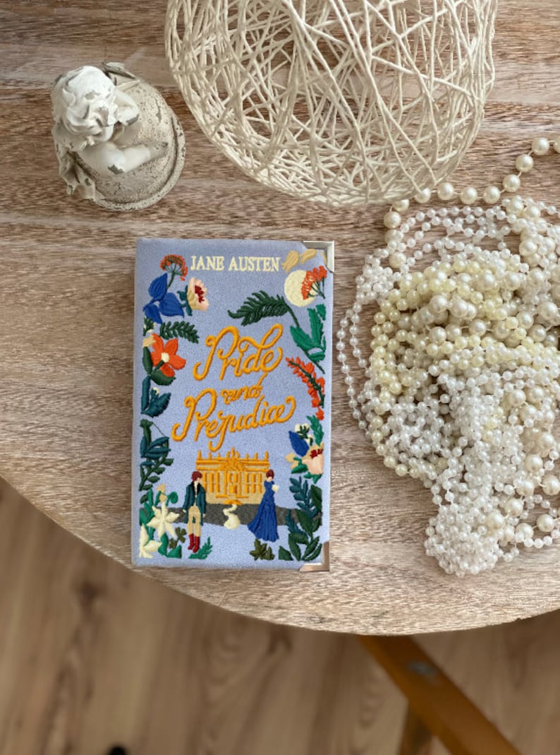 May include: A blue book cover with the title "Pride and Prejudice" embroidered in yellow and orange thread. The cover also features a small embroidered illustration of a building and two figures.
