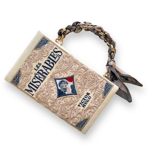 May include: A white book clutch bag with a gold chain strap and a brown and gray silk scarf. The bag is embroidered with the title "Les Miserables" and the author "Victor Hugo".