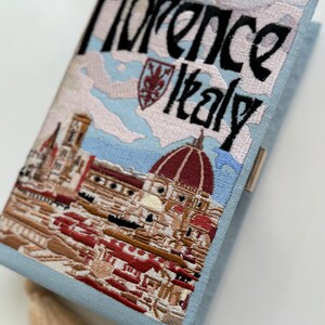 Pu&ograve; includere: Una pochette ricamata di colore azzurro chiaro con una scena di Firenze, Italia. La borsa presenta il Duomo, un paesaggio urbano e il testo "Florence Italy". La borsa ha un fiocco.