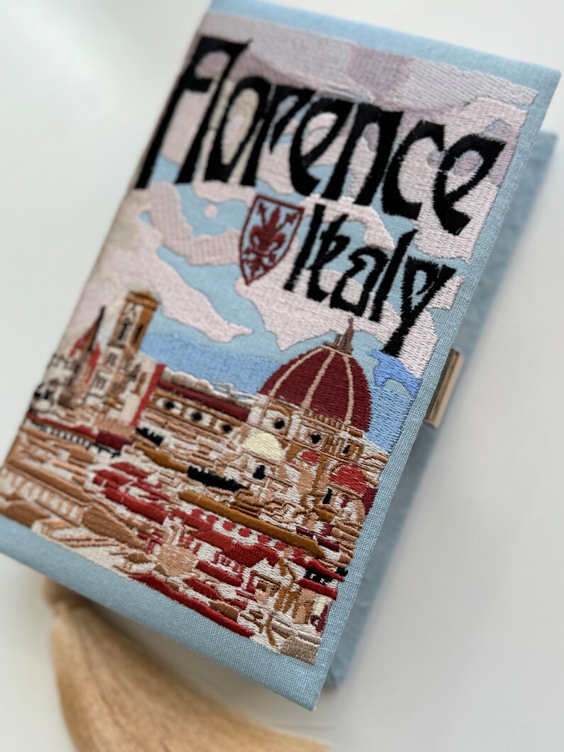 Pu&ograve; includere: Una pochette ricamata di colore azzurro chiaro con una scena di Firenze, Italia. La borsa presenta il Duomo, un paesaggio urbano e il testo "Florence Italy". La borsa ha un fiocco.