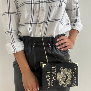 K&ouml;nnte beinhalten: Ein schwarzer Lederrock mit einer goldenen Kettenriementasche. Die Tasche ist in Form eines Buches mit dem Titel "Die Kunst des Krieges" und dem Autor "Sun Tzu" in goldenen Lettern gedruckt. Die Tasche hat ein goldenes Drachenmotiv.