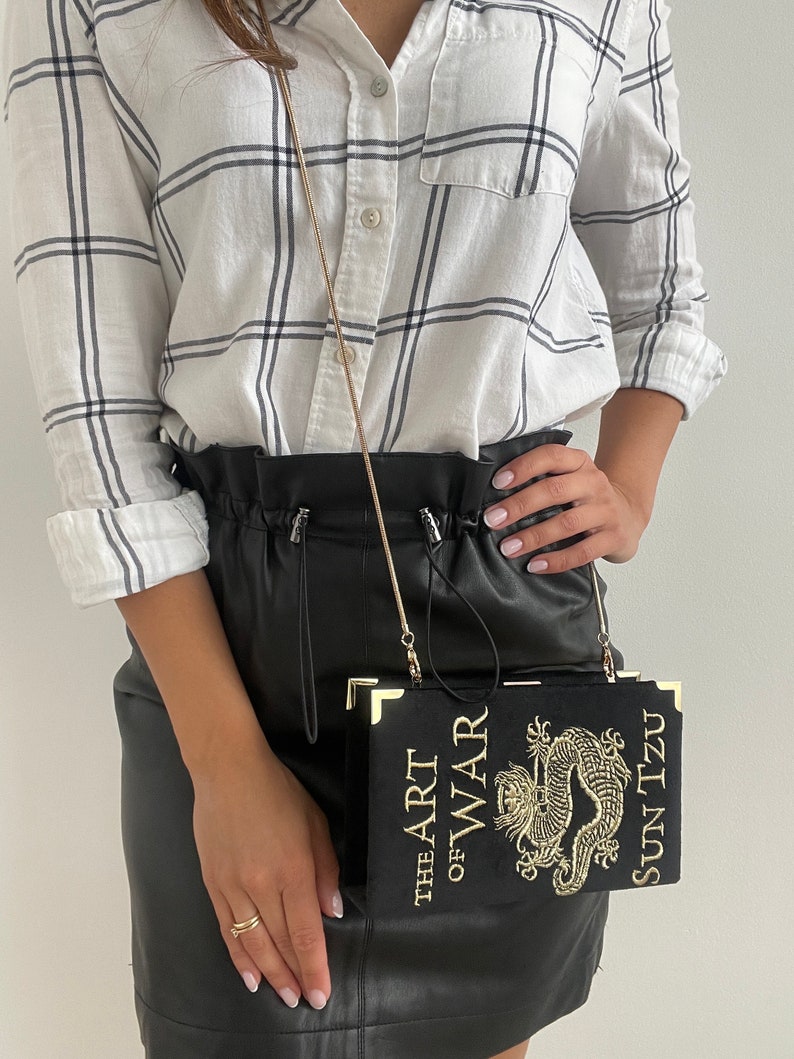 K&ouml;nnte beinhalten: Ein schwarzer Lederrock mit einer goldenen Kettenriementasche. Die Tasche ist in Form eines Buches mit dem Titel "Die Kunst des Krieges" und dem Autor "Sun Tzu" in goldenen Lettern gedruckt. Die Tasche hat ein goldenes Drachenmotiv.