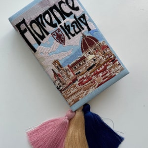 Pu&ograve; includere: Una pochette ricamata di colore azzurro chiaro con una scena di Firenze, Italia, e il testo "Florence Italy". La pochette ha tre nappine di colore rosa, beige e blu scuro.