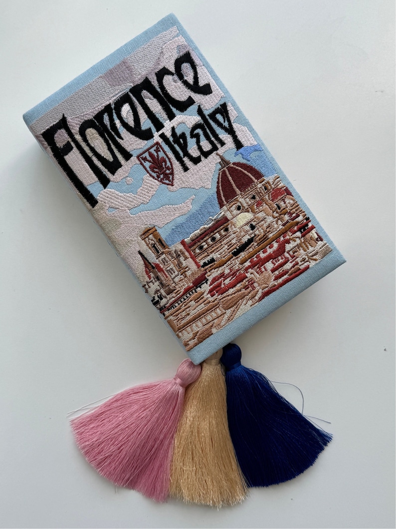 Pu&ograve; includere: Una pochette ricamata di colore azzurro chiaro con una scena di Firenze, Italia, e il testo "Florence Italy". La pochette ha tre nappine di colore rosa, beige e blu scuro.