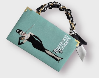 May include: A teal blue clutch bag with a black and white braided strap and gold accents. The bag features an embroidered image of Audrey Hepburn from the movie "Breakfast at Tiffany's".