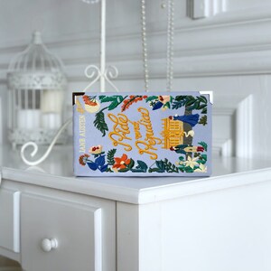 May include: A light blue book with the title "Pride and Prejudice" embroidered in yellow lettering. The book cover features a floral design with a small embroidered image of a building.