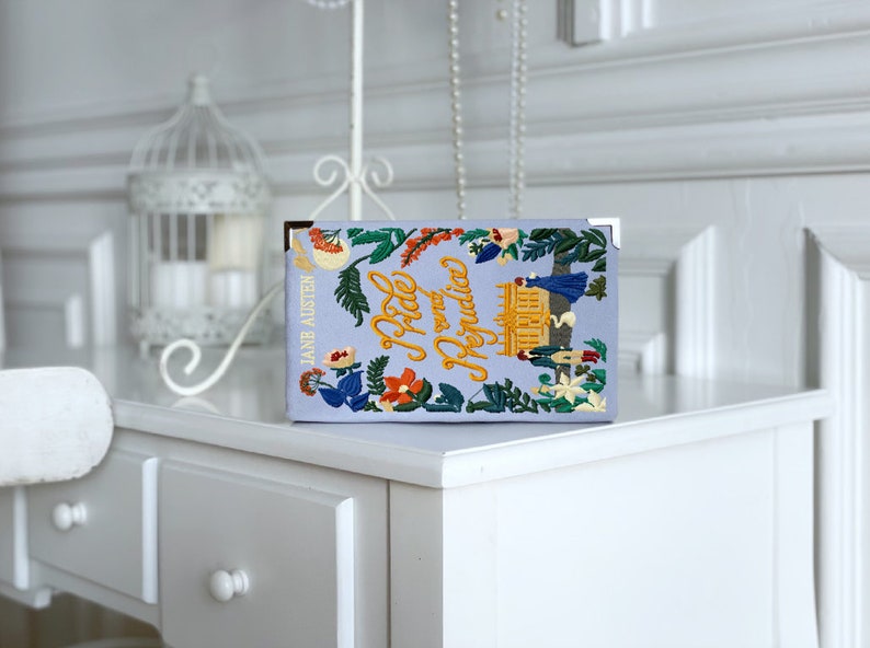 May include: A light blue book with the title "Pride and Prejudice" embroidered in yellow lettering. The book cover features a floral design with a small embroidered image of a building.