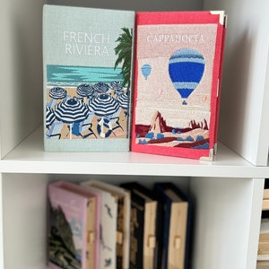 May include: Two decorative books with embroidered covers. The book on the left has a blue and white beach scene with the text "French Riviera". The book on the right has a red and white hot air balloon scene with the text "Cappadocia".