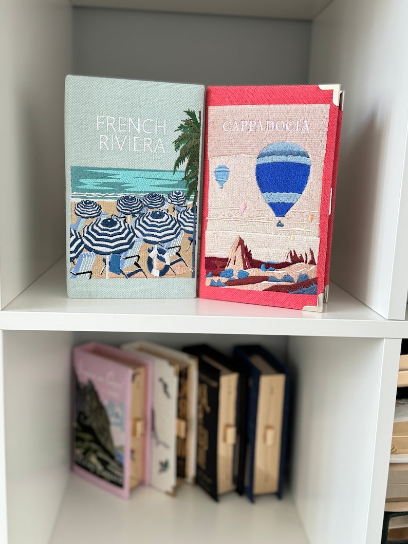 May include: Two decorative books with embroidered covers. The book on the left has a blue and white beach scene with the text "French Riviera". The book on the right has a red and white hot air balloon scene with the text "Cappadocia".